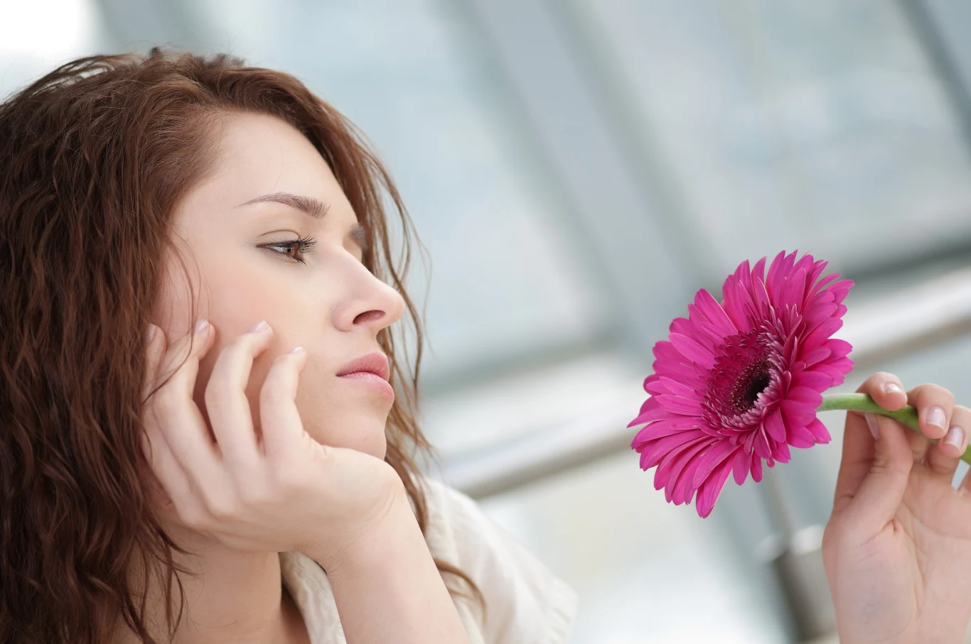 Młoda kobieta z długimi, brązowymi włosami wpatruje się zamyślona w różowy kwiat gerbery trzymany w dłoni.