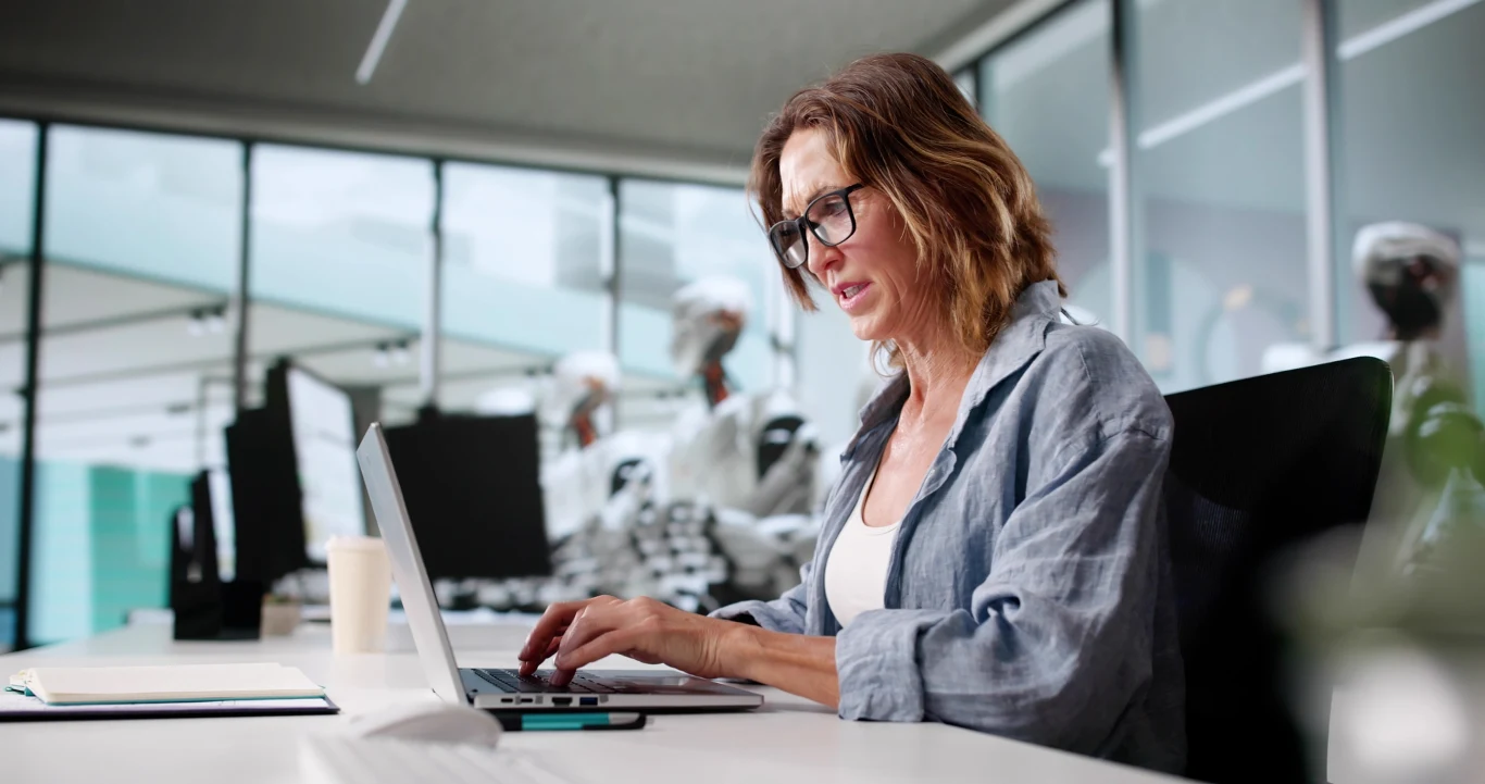 Kobieta w okularach pracuje przy laptopie w nowoczesnym biurze, w tle widoczne są roboty humanoidalne wykonujące różne czynności przy biurkach.