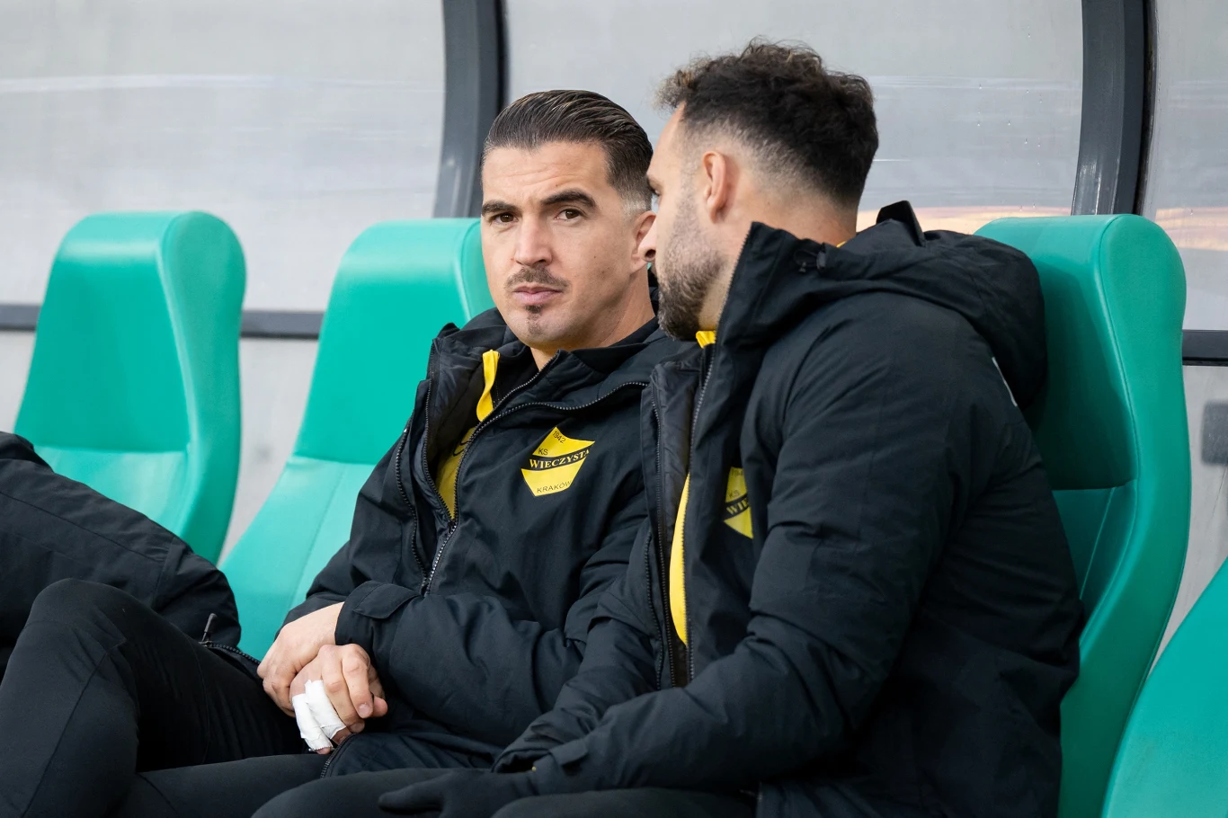 Dwóch mężczyzn w sportowych kurtkach z emblematem klubu siedzi obok siebie na zielonych plastikowych krzesłach, jeden z nich ma zabandażowaną dłoń i patrzy przed siebie, drugi zwrócony jest w jego stronę i prowadzi rozmowę.