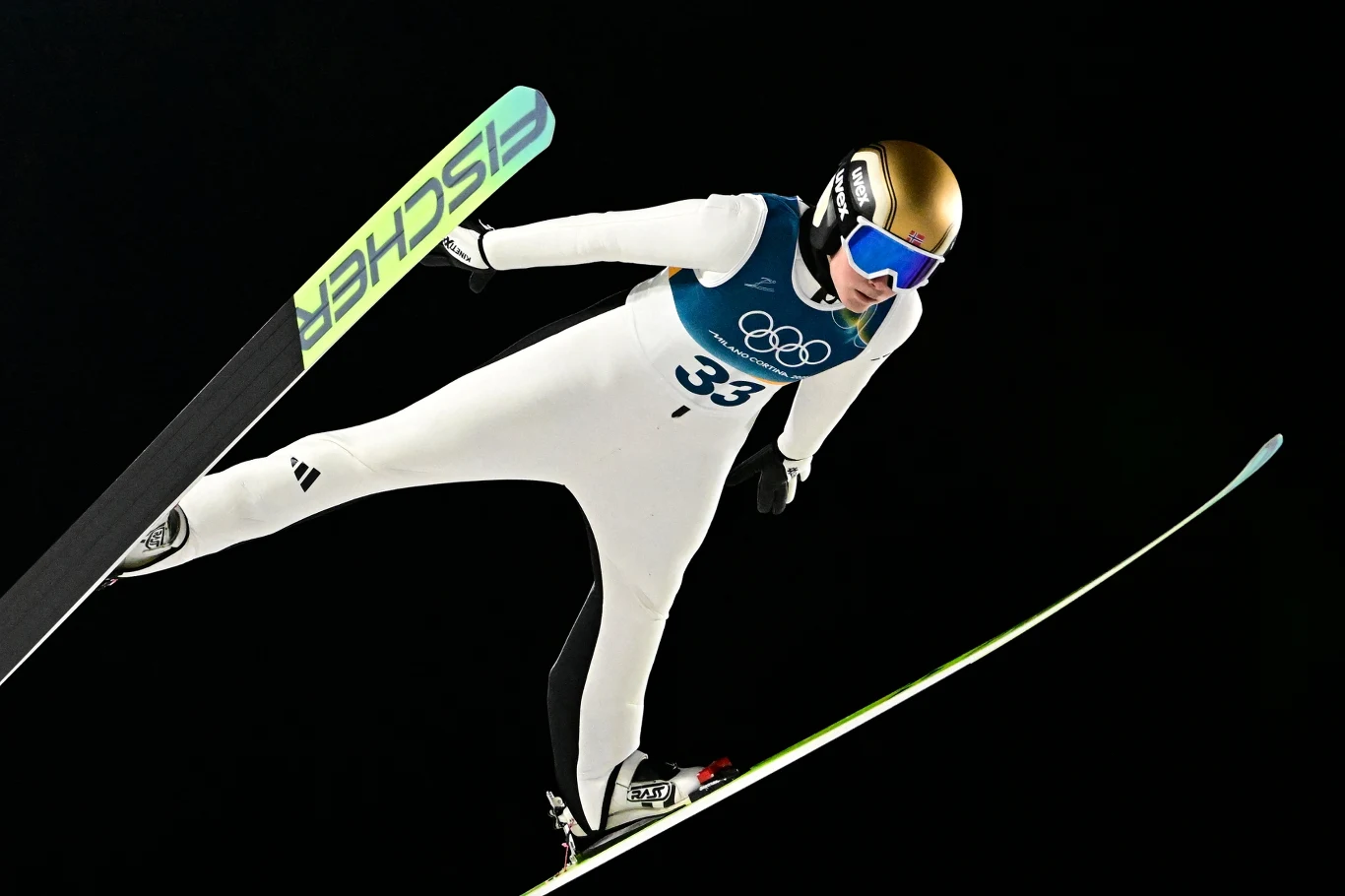 Silje Opseth podczas igrzysk olimpijskich Silje Opseth podczas igrzysk olimpijskich, w biało-niebieskim stroju i złotym kasku, w locie na tle ciemnego nieba.