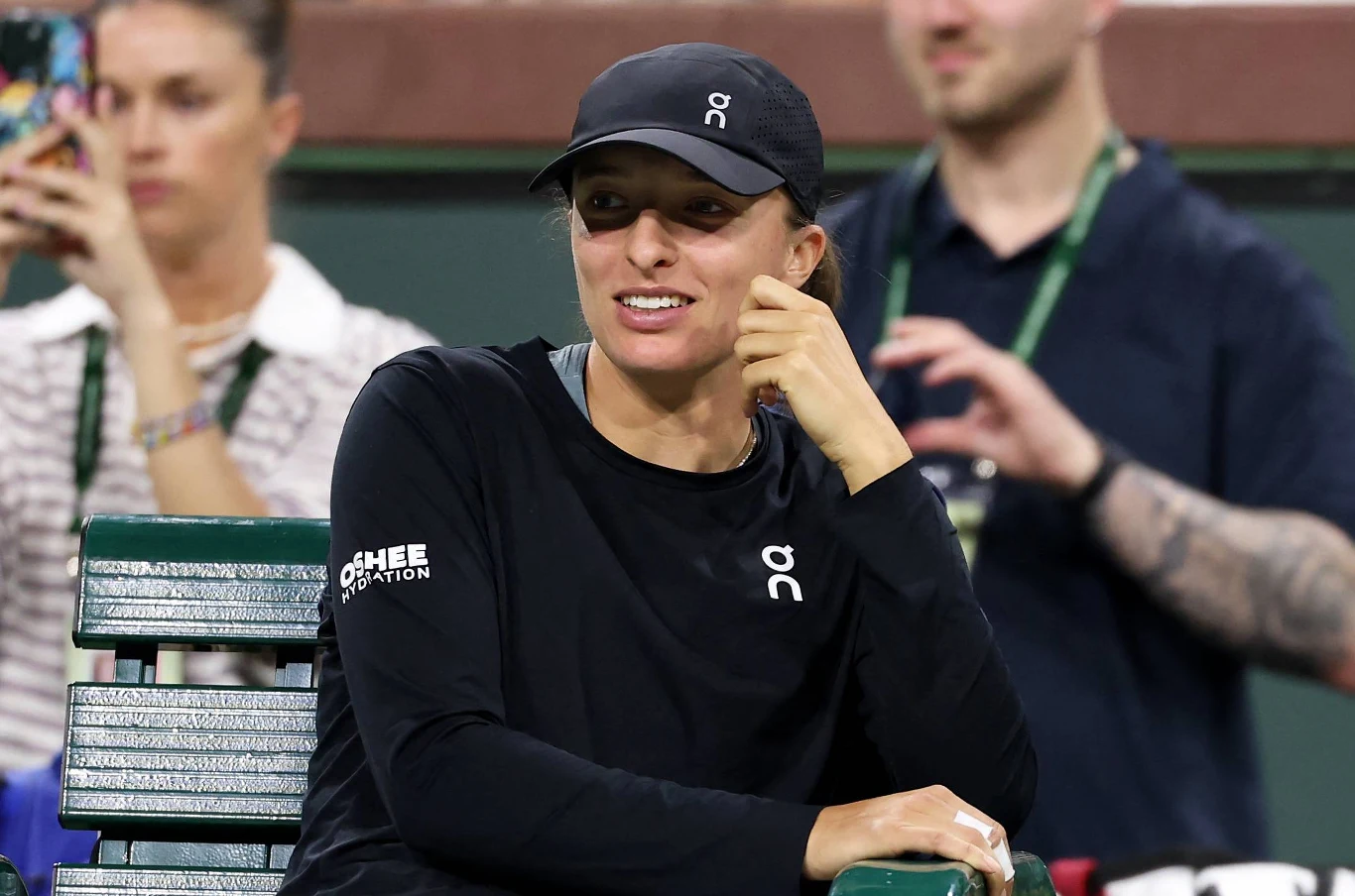 Una mujer con gorra deportiva negra y sudadera con el logo de la marca ON, sentada en un banco de tenis, sonriendo, con figuras borrosas de otras personas al fondo.