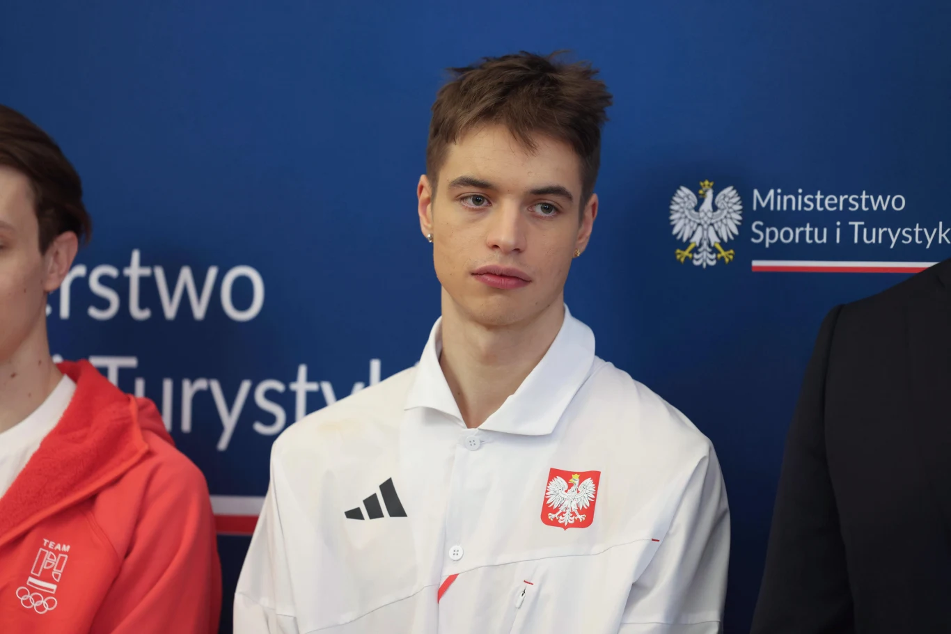 Młody mężczyzna w białej koszulce z godłem Polski stoi na tle niebieskiej ściany z logotypem Ministerstwa Sportu i Turystyki, obok osoby w czerwonej bluzie oraz osoby w ciemnym garniturze.