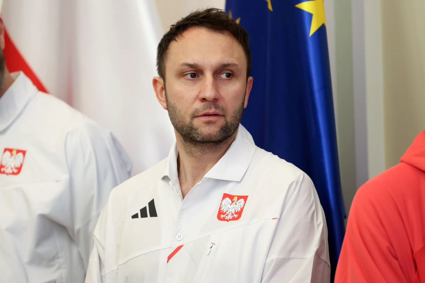 Mężczyzna w białej koszuli z godłem Polski i logo sportowym stoi na tle biało-czerwonej oraz niebieskiej flagi z gwiazdami, patrząc na bok z poważnym wyrazem twarzy.