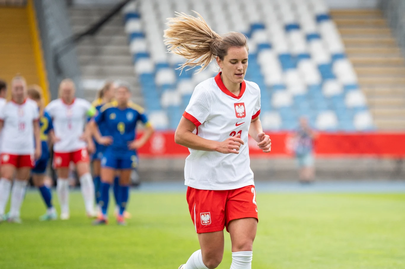 Zawodniczka reprezentacji Polski w piłce nożnej w trakcie meczu, ubrana w biało-czerwony strój, biegnie po murawie, w tle zawodniczki drużyny przeciwnej w niebieskich strojach oraz puste trybuny stadionu.