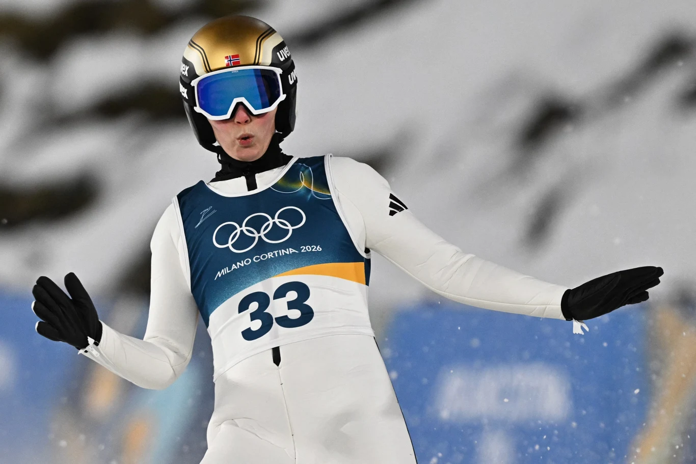 Silje Opseth Zawodnik w białym kombinezonie narciarskim z numerem 33 i kaskiem w odcieniach złota podczas skoku narciarskiego, w tle rozmyty krajobraz górski.