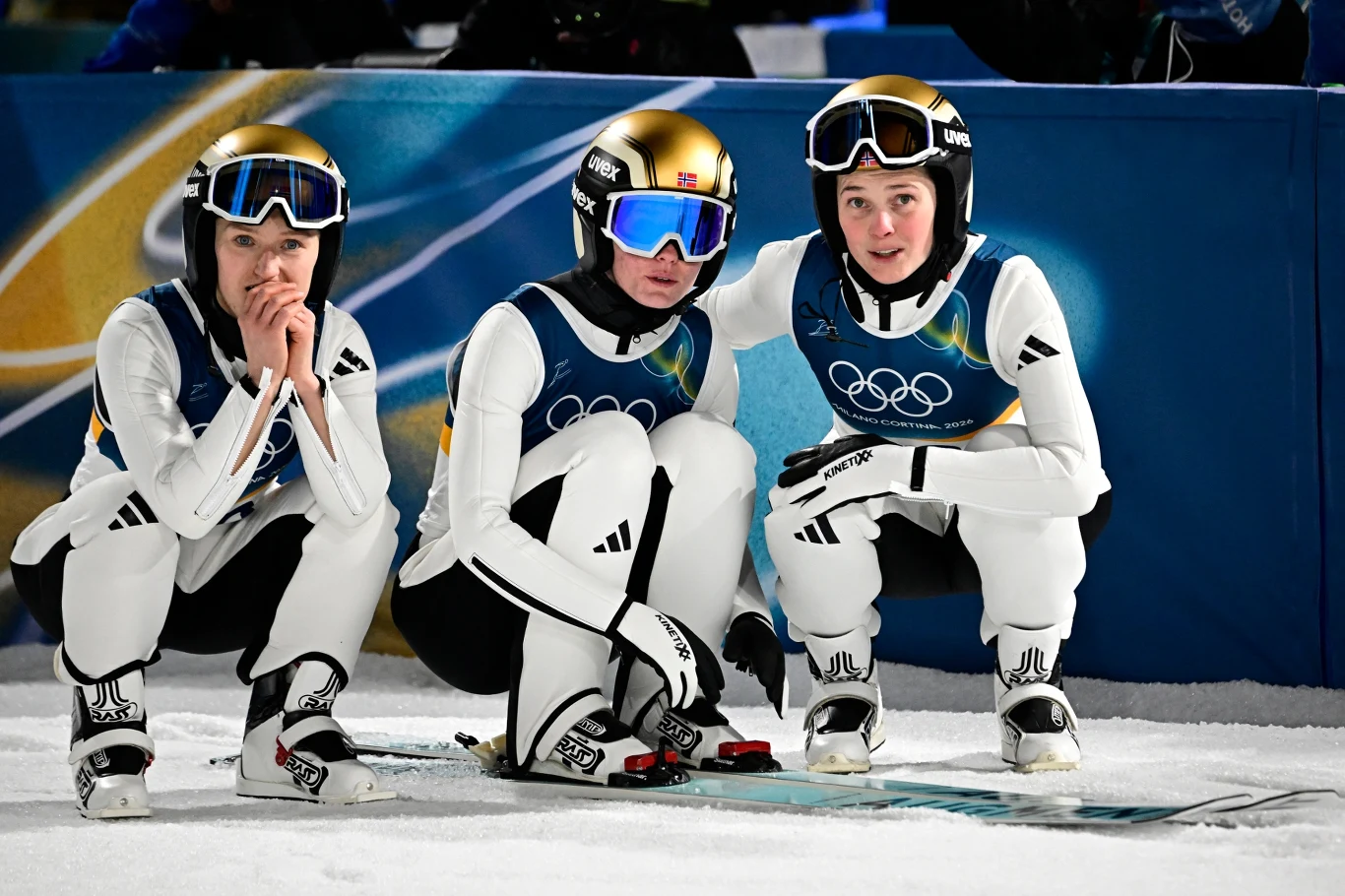 Norweskie skoczkinie narciarskie Trzy narciarki ubrane w białe kombinezony i złote kaski w skupieniu kucają obok siebie na śniegu przy torze podczas zawodów olimpijskich, zwracając uwagę na to, co dzieje się na stoku.