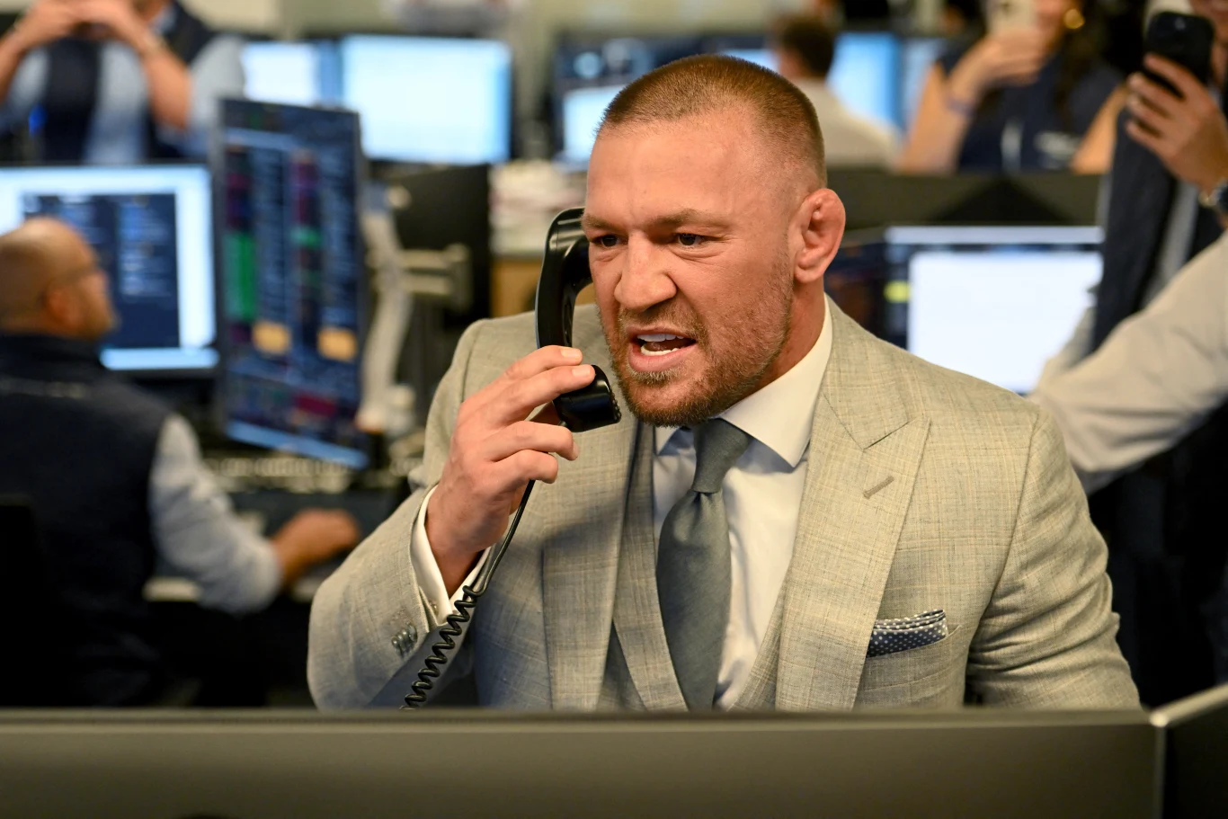 Conor Mcgregor Conor McGregor w garniturze rozmawia przez telefon przy biurku z monitorami giełdowymi i innymi pracownikami.
