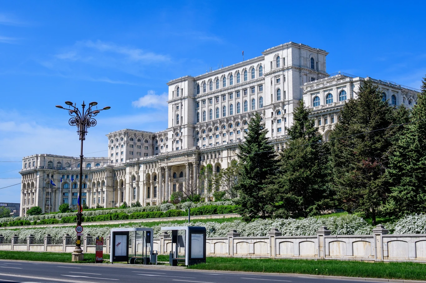 Monumentalny budynek o klasycystycznej architekturze z licznymi kolumnami i oknami, otoczony zielenią i drzewami iglastymi; na pierwszym planie widać ulicę oraz przystanek autobusowy z wiatami.