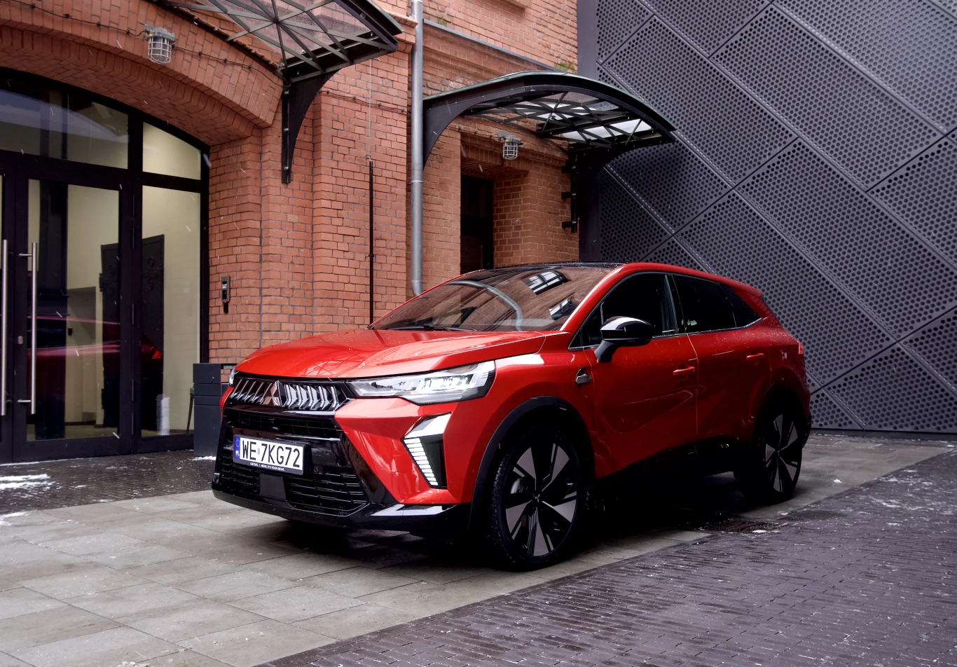 Czerwony nowoczesny samochód SUV zaparkowany przed budynkiem z czerwonej cegły, z ciemnymi drzwiami wejściowymi i metalowym daszkiem nad drzwiami.