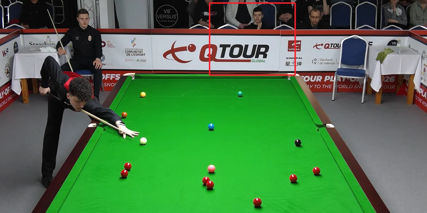 Zawodnik wykonuje precyzyjny strzał przy stole bilardowym snookerowym, otoczony kolorowymi bilami i obserwowany przez sędziego oraz widownię w tle; dominują jasne, turniejowe barwy oraz profesjonalna atmosfera rozgrywek.