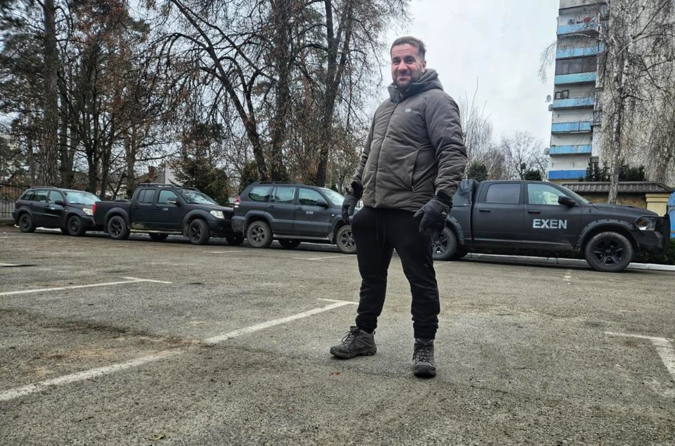 Mężczyzna w zimowej kurtce i rękawiczkach stoi na pustym parkingu, za nim rząd ciemnych samochodów i budynek mieszkalny w tle, w otoczeniu nagich drzew.