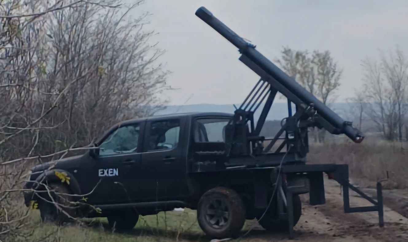Czarny samochód terenowy z zamontowaną na pace wyrzutnią rakiet w pozycji gotowej do wystrzału, otoczony przez suche krzewy i drzewa na nieutwardzonej drodze.