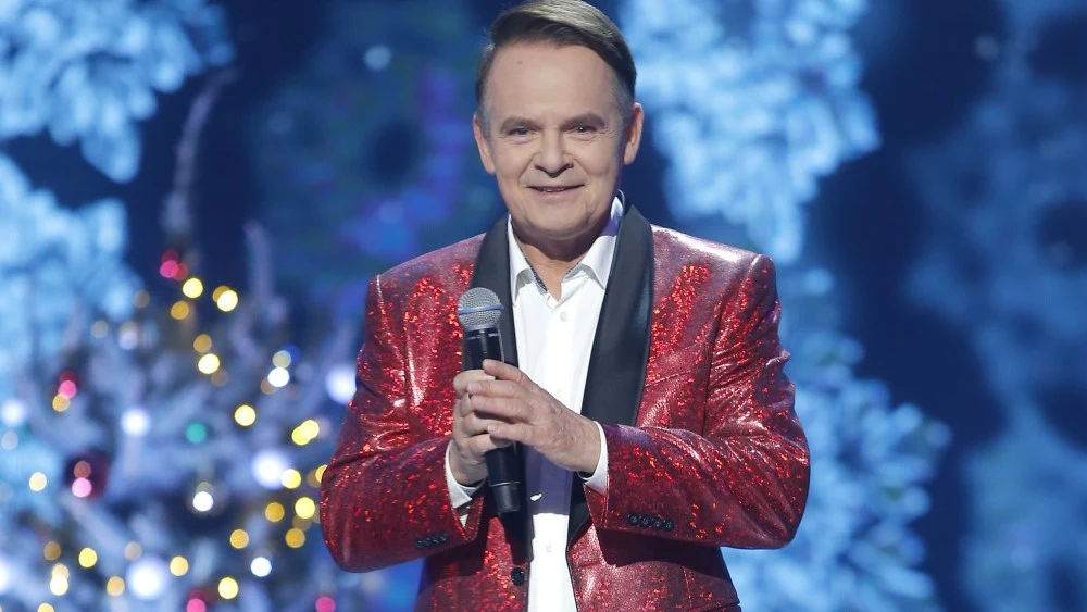 Piotr Pręgowski Piotr Pręgowski w czerwonej, błyszczącej marynarce z mikrofonem na scenie, w tle choinka i światełka.