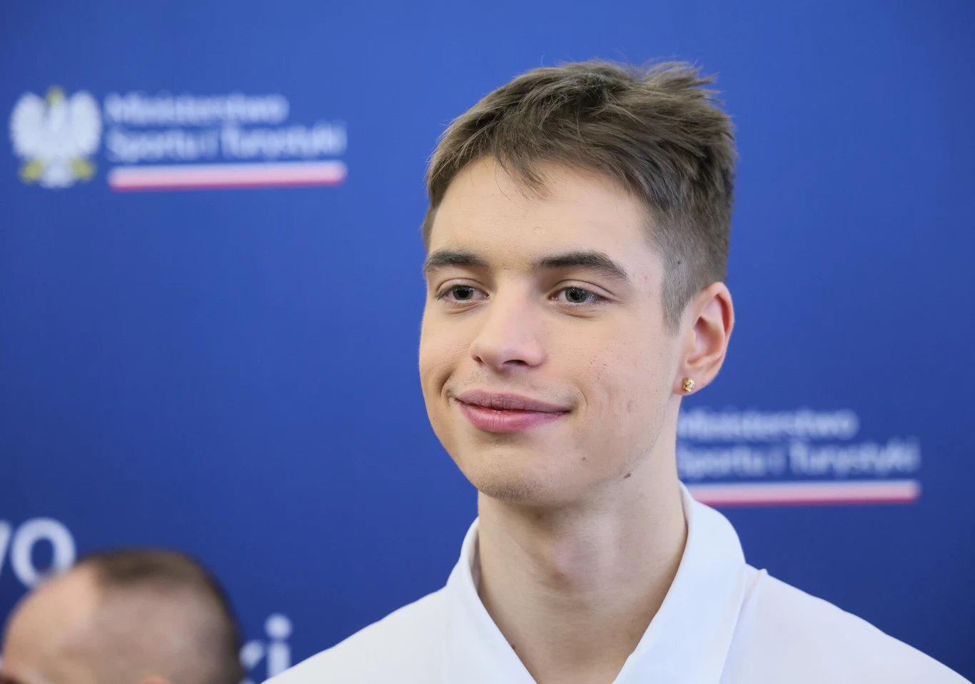 Młody mężczyzna z krótkimi brązowymi włosami uśmiecha się lekko, ubrany w białą koszulę, na tle niebieskiej planszy z logotypem Ministerstwa Sportu i Turystyki.