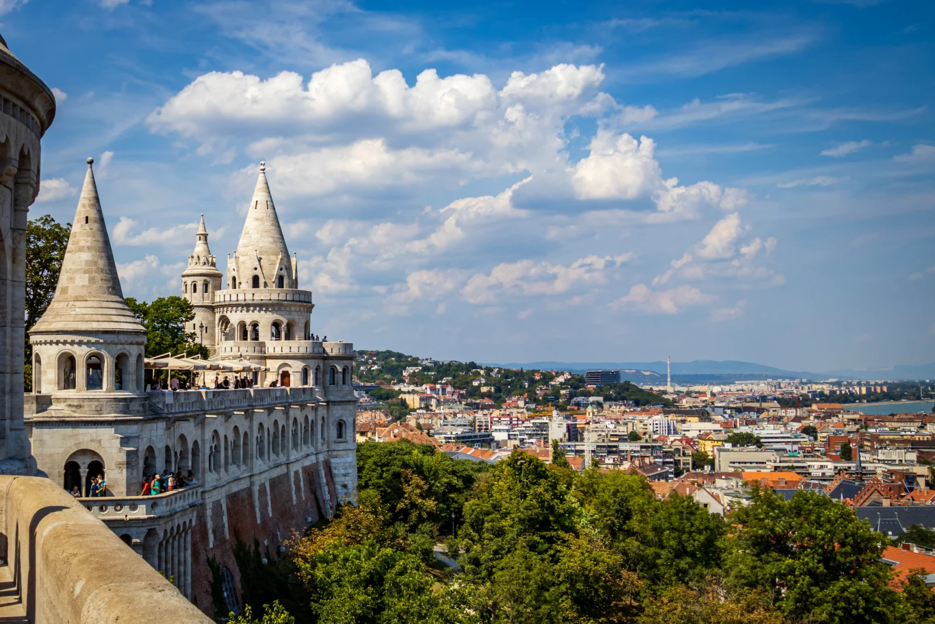 Baszta Rybacka z białymi, strzelistymi wieżami i tarasami góruje nad panoramą Budapesztu, otoczona zielenią drzew i rozległym widokiem na miasto oraz Dunaj w słoneczny dzień.