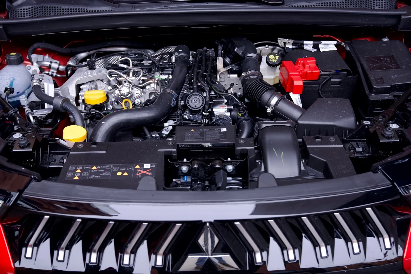 Silnik samochodowy z widocznymi przewodami, paskami, akumulatorem i różnymi złączami technicznymi pod maską pojazdu, z dominacją czarnych, srebrnych i żółtych elementów.