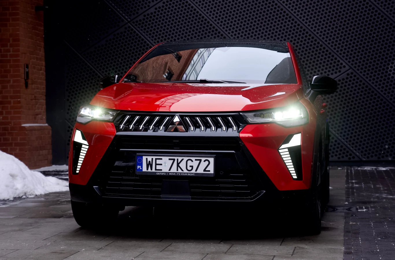 Czerwony samochód Mitsubishi z polską tablicą rejestracyjną, nowoczesnym zderzakiem i charakterystycznym układem świateł LED, ustawiony na brukowanej nawierzchni obok ceglanej ściany.