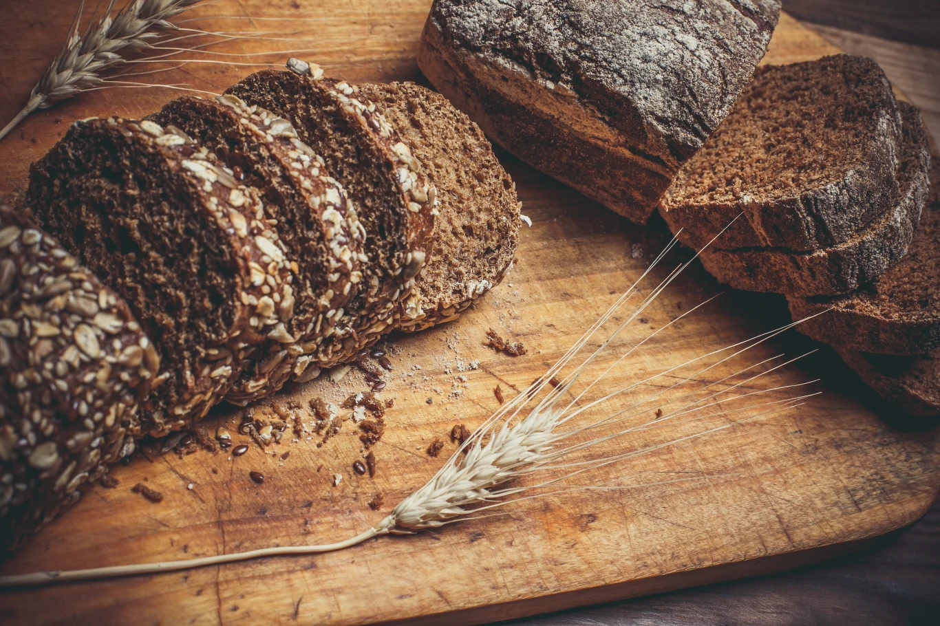Pokrojony ciemny chleb z ziarnami leży na drewnianej desce obok całego bochenka i pojedynczych kłosów zboża.