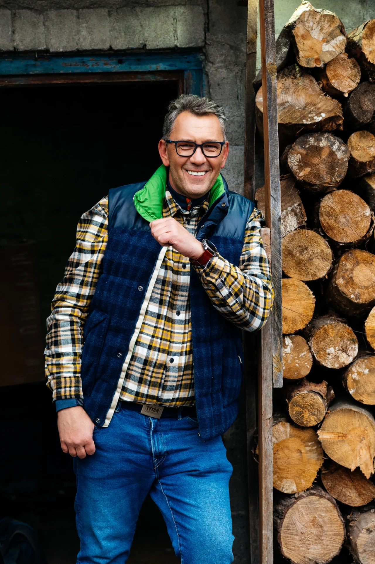 Mężczyzna w okularach ubrany w kraciastą koszulę, niebieską kamizelkę i dżinsy stoi obok stosu drewna opałowego, uśmiechając się szeroko.