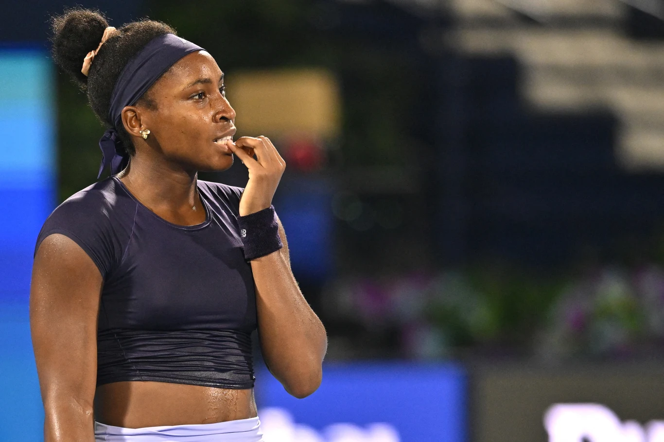 Młoda tenisistka w sportowym stroju z opaską na głowie stoi na korcie, patrząc zamyślona w bok i trzymając palec przy ustach, wyrażając skupienie lub niepewność.