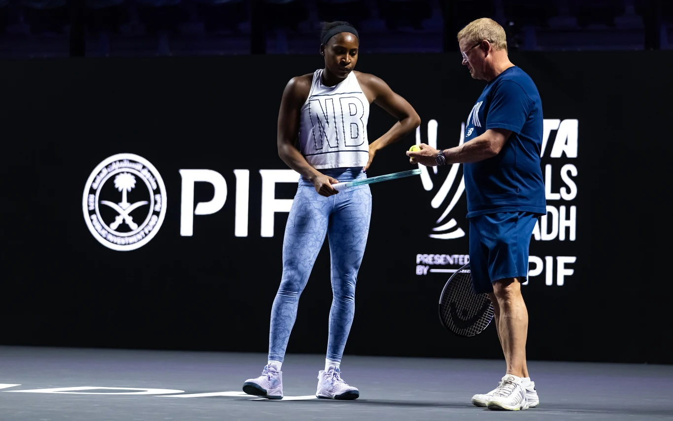 Tenisistka w jasnym stroju sportowym i trener w granatowym ubraniu stoją razem na korcie, wymieniając się rakietą i piłką na tle ciemnego ekranu z logotypami turnieju WTA oraz napisem PIF.