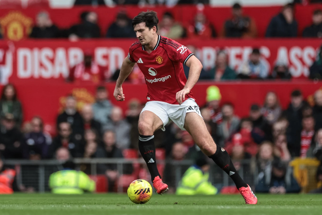 Piłkarz Manchesteru United w czerwonej koszulce i białych spodenkach prowadzi żółto-czerwoną piłkę podczas meczu na stadionie, w tle widownia i zabezpieczenie stadionu.