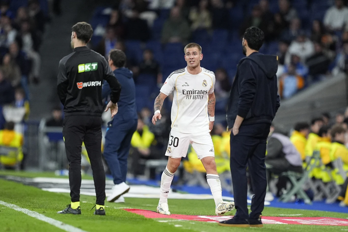 Piłkarz w białym stroju Realu Madryt stoi przy linii bocznej boiska podczas meczu, wokół niego trenerzy i sztab szkoleniowy, w tle ławka rezerwowych oraz żółto ubrani porządkowi na trybunach.