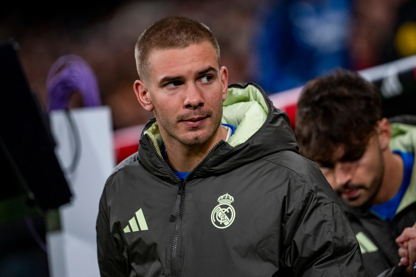 Młody mężczyzna w sportowej kurtce z herbem Realu Madryt, stojący na tle sprzętu technicznego i innych członków drużyny.