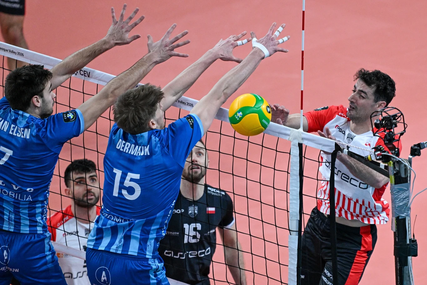 Zawodnik w biało-czerwonym stroju atakuje piłkę przy siatce, podczas gdy dwóch siatkarzy w niebieskich koszulkach próbuje zablokować jego zagranie, a w tle widoczni są pozostali gracze obserwujący akcję.