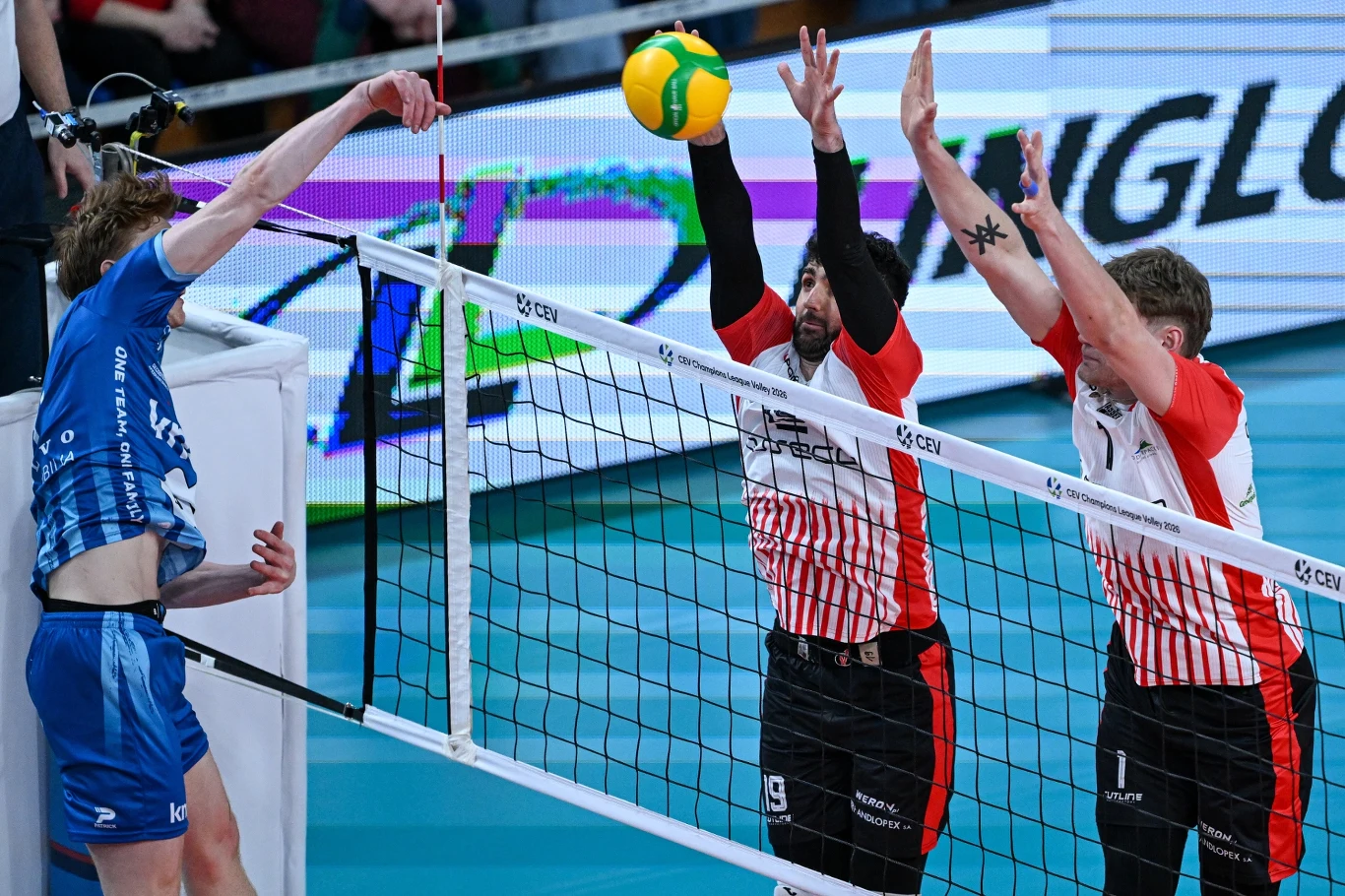 Dwóch siatkarzy w białych koszulkach z czerwonymi paskami wykonuje blok przy siatce, skutecznie zatrzymując atakującego zawodnika w niebieskim stroju; żółto-zielona piłka znajduje się tuż nad siatką.