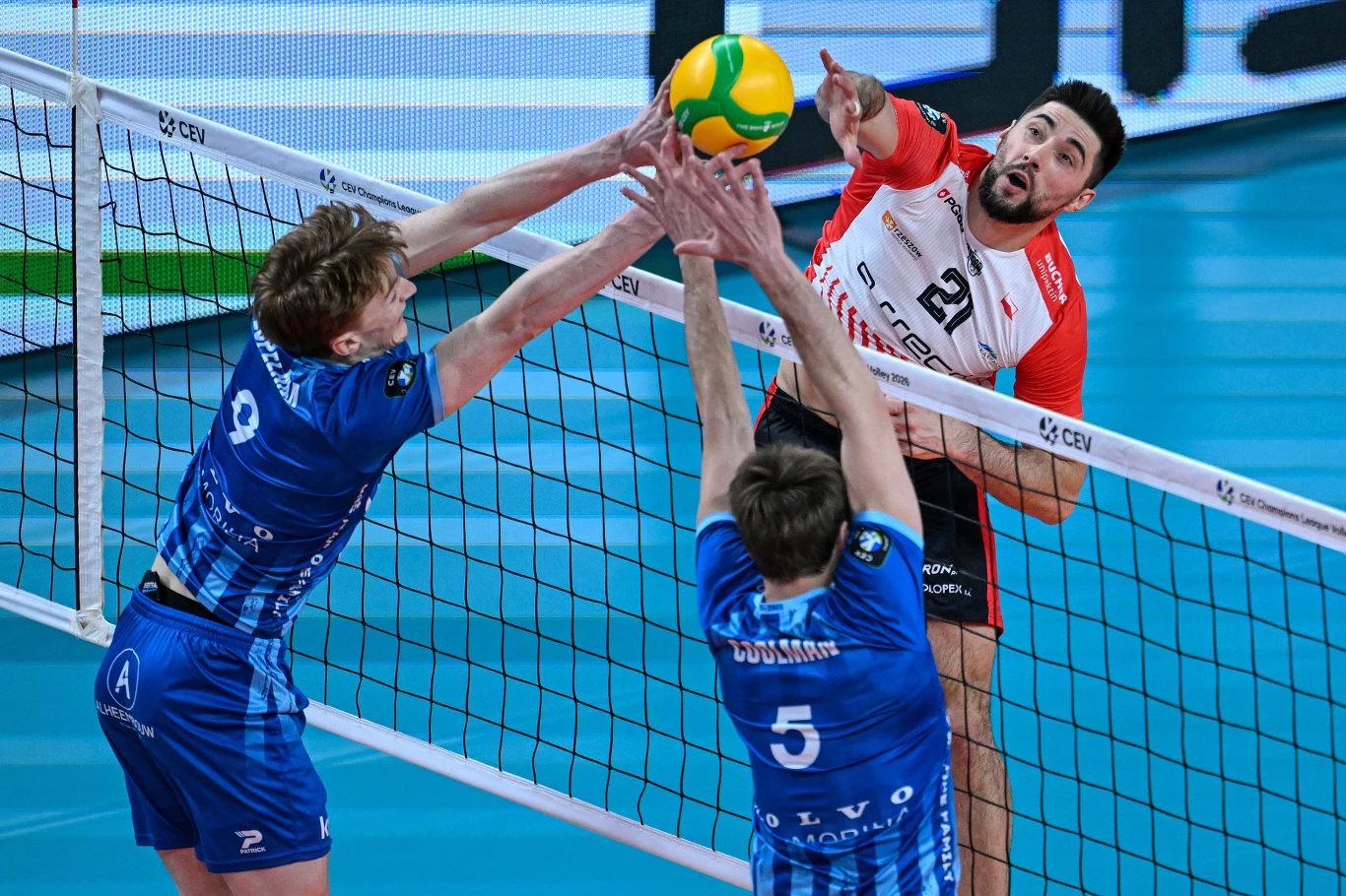 Zawodnik w czerwono-białym stroju atakuje piłkę siatkarską tuż nad siatką, podczas gdy dwóch graczy w niebieskich strojach próbuje zablokować atak, wyciągając ręce w górę.