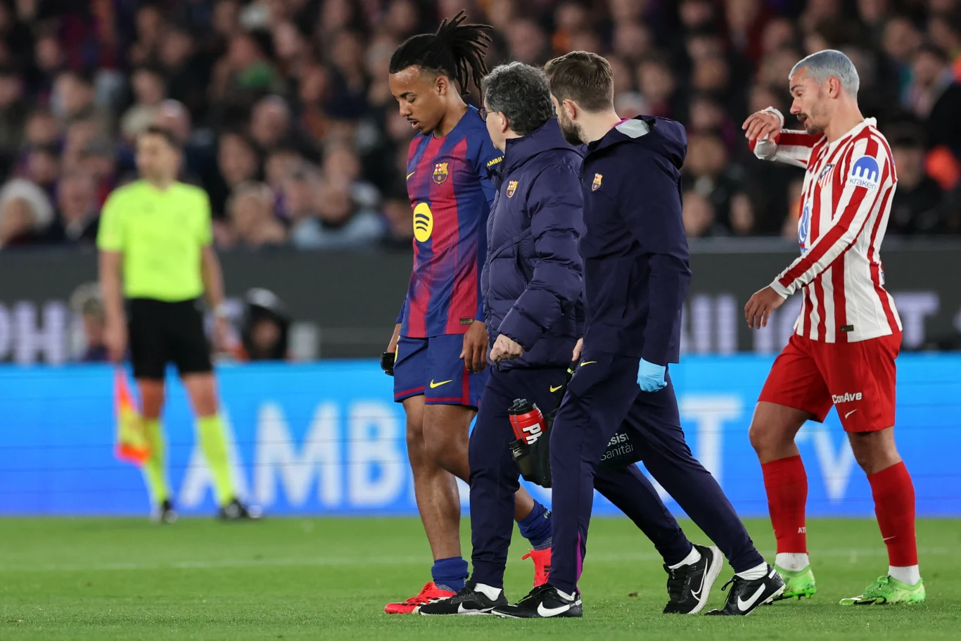 Piłkarz FC Barcelony opuszcza boisko w towarzystwie sztabu medycznego, podczas gdy zawodnik drużyny przeciwnej w biało-czerwonych paskach obserwuje sytuację.