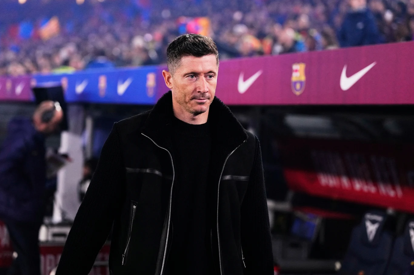 Mężczyzna z krótkimi ciemnymi włosami ubrany w czarną kurtkę stoi na tle ławki rezerwowych na stadionie piłkarskim z widocznymi logotypami FC Barcelony i Nike, w otoczeniu rozświetlonej trybuny pełnej kibiców.