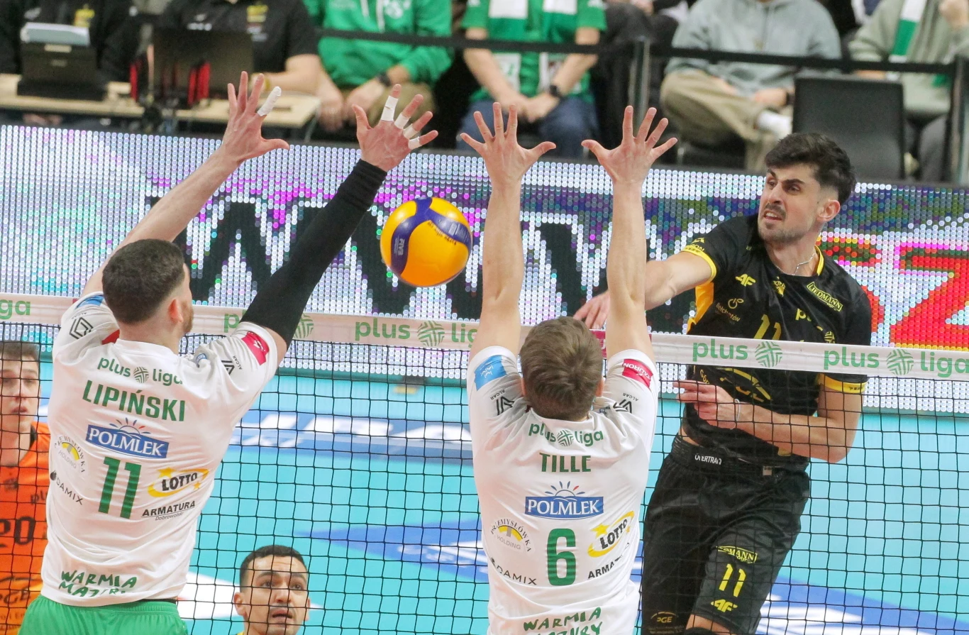 Trzech siatkarzy rywalizuje przy siatce, dwóch zawodników w białych koszulkach próbuje zablokować atakującego w czarno-żółtym stroju, nad siatką unosi się piłka do siatkówki.