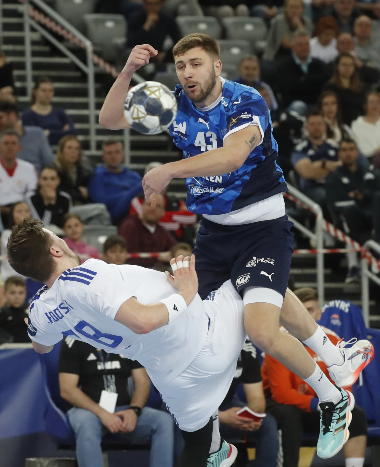 Dwóch piłkarzy ręcznych w dynamicznym starciu podczas meczu; zawodnik w niebieskim stroju unosi się nad ziemią w momencie rzutu, a rywal w białym stroju próbuje go zatrzymać, podczas gdy piłka znajduje się w powietrzu tuż obok nich, a w tle widoczna je...