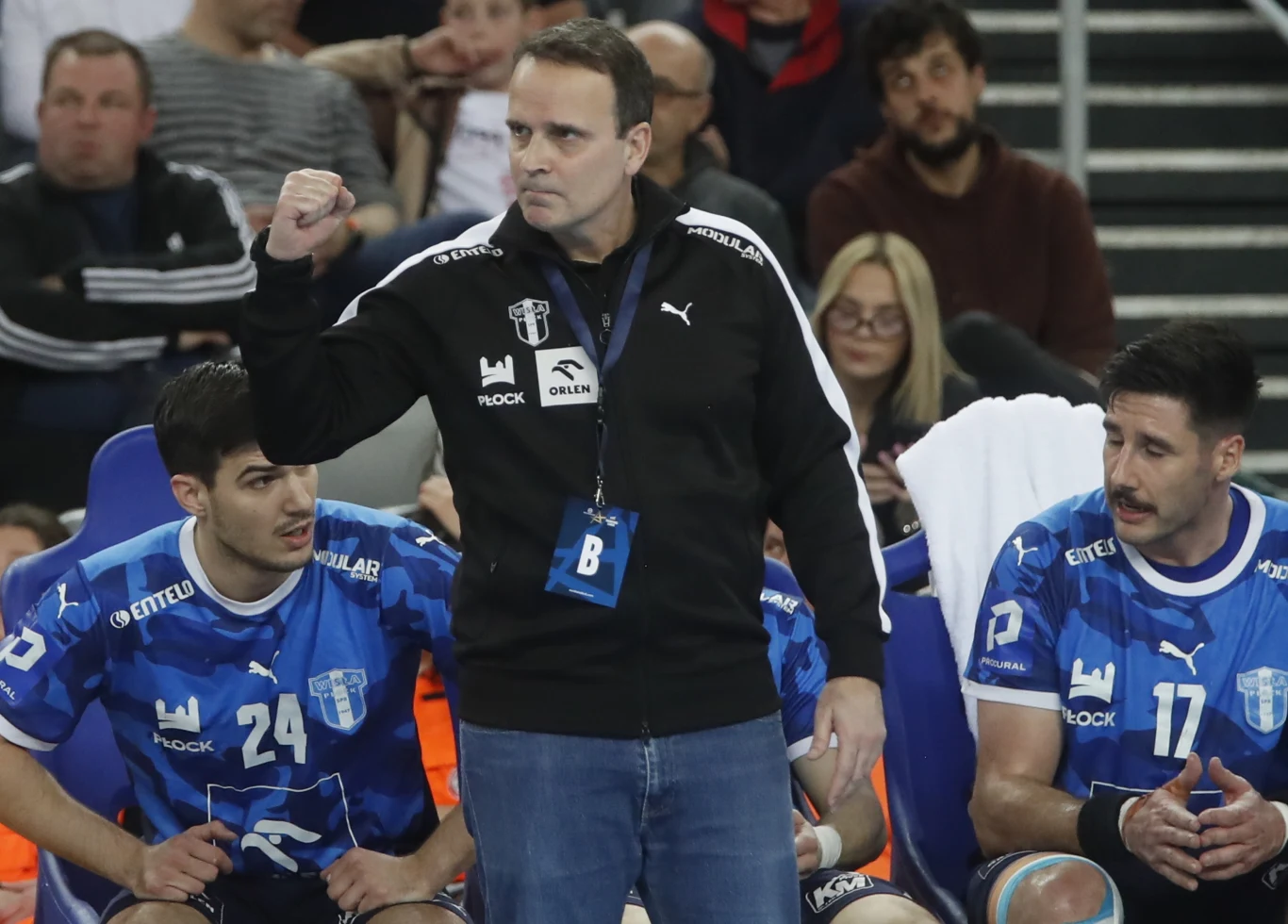 Trener ubrany w czarną bluzę z zaciśniętą pięścią stoi między dwoma zawodnikami drużyny piłki ręcznej w niebieskich koszulkach z numerami 24 i 11, którzy siedzą na ławce rezerwowych podczas meczu.