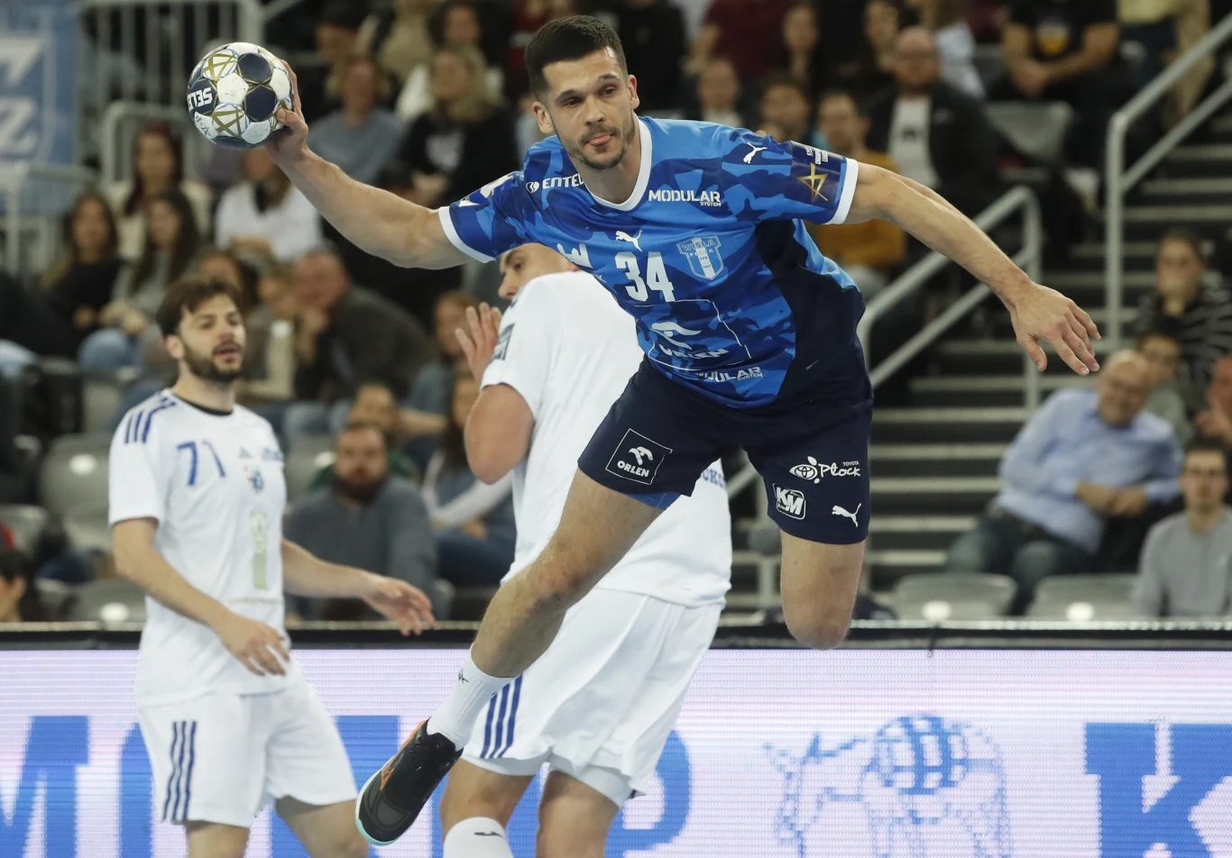 Zawodnik w niebieskiej koszulce podczas dynamicznego rzutu piłką ręczną w powietrzu, otoczony przez przeciwnika w białym stroju, w tle widownia na trybunach.