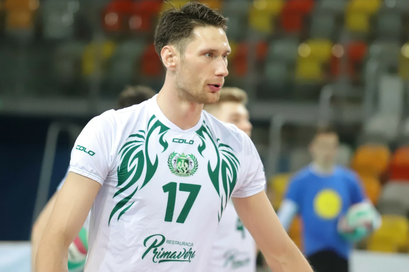 Siatkarz w białej koszulce z numerem 17 i zielonymi elementami graficznymi na tle hali sportowej, wokół widoczni inni zawodnicy i trybuny z kolorowymi miejscami siedzącymi.