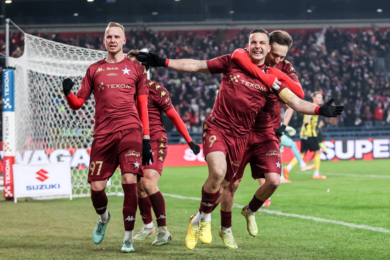 Piłkarze drużyny w bordowych strojach świętują zdobycie gola na stadionie, jeden zawodnik uśmiechnięty z rozłożonymi ramionami, a inny wskakuje mu na plecy, w tle widać trybuny pełne kibiców.