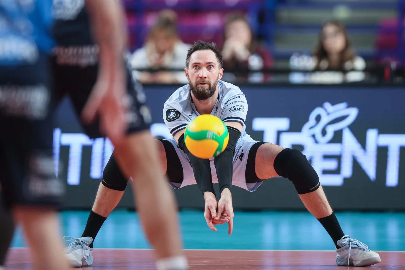 Siatkarz w białej koszulce z kolanami zgiętymi w szerokim rozkroku wykonuje odbiór niskiej piłki do siatkówki, skupiony na precyzyjnym kontakcie z piłką podczas meczu na hali sportowej.