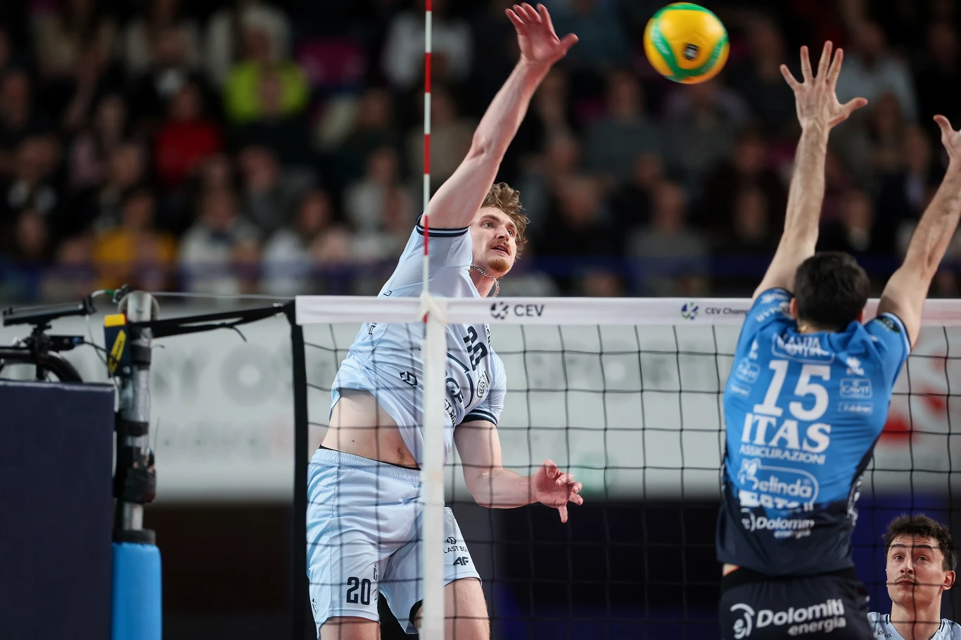 Dwóch siatkarzy w akcji przy siatce, jeden wykonuje atak, drugi próbuje blokować piłkę; dynamiczny moment meczu siatkarskiego na tle publiczności na trybunach.