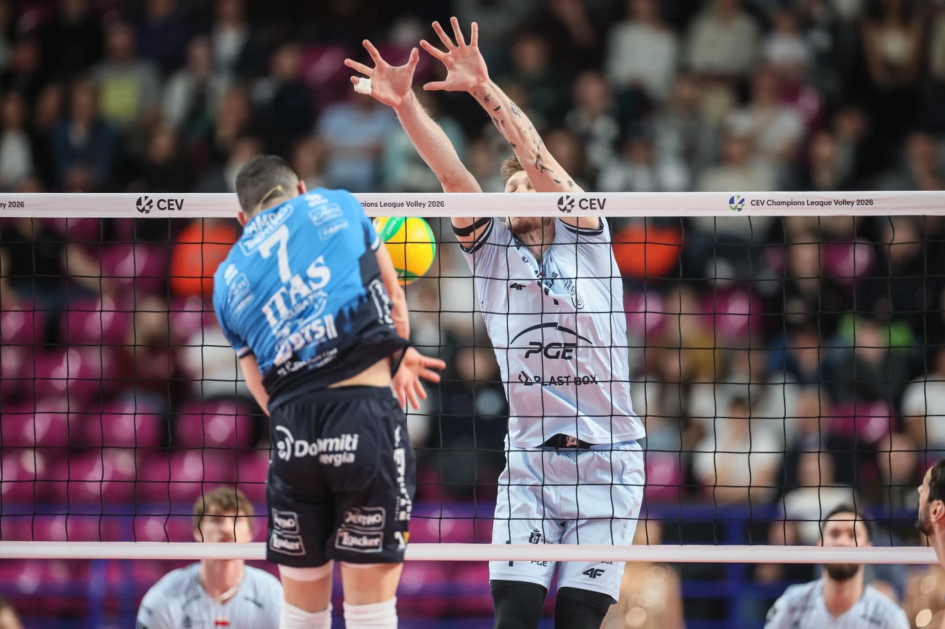 Dwóch siatkarzy w trakcie dynamicznej akcji przy siatce, jeden z nich wykonuje blok, drugi przygotowuje się do ataku w trakcie meczu siatkówki halowej.