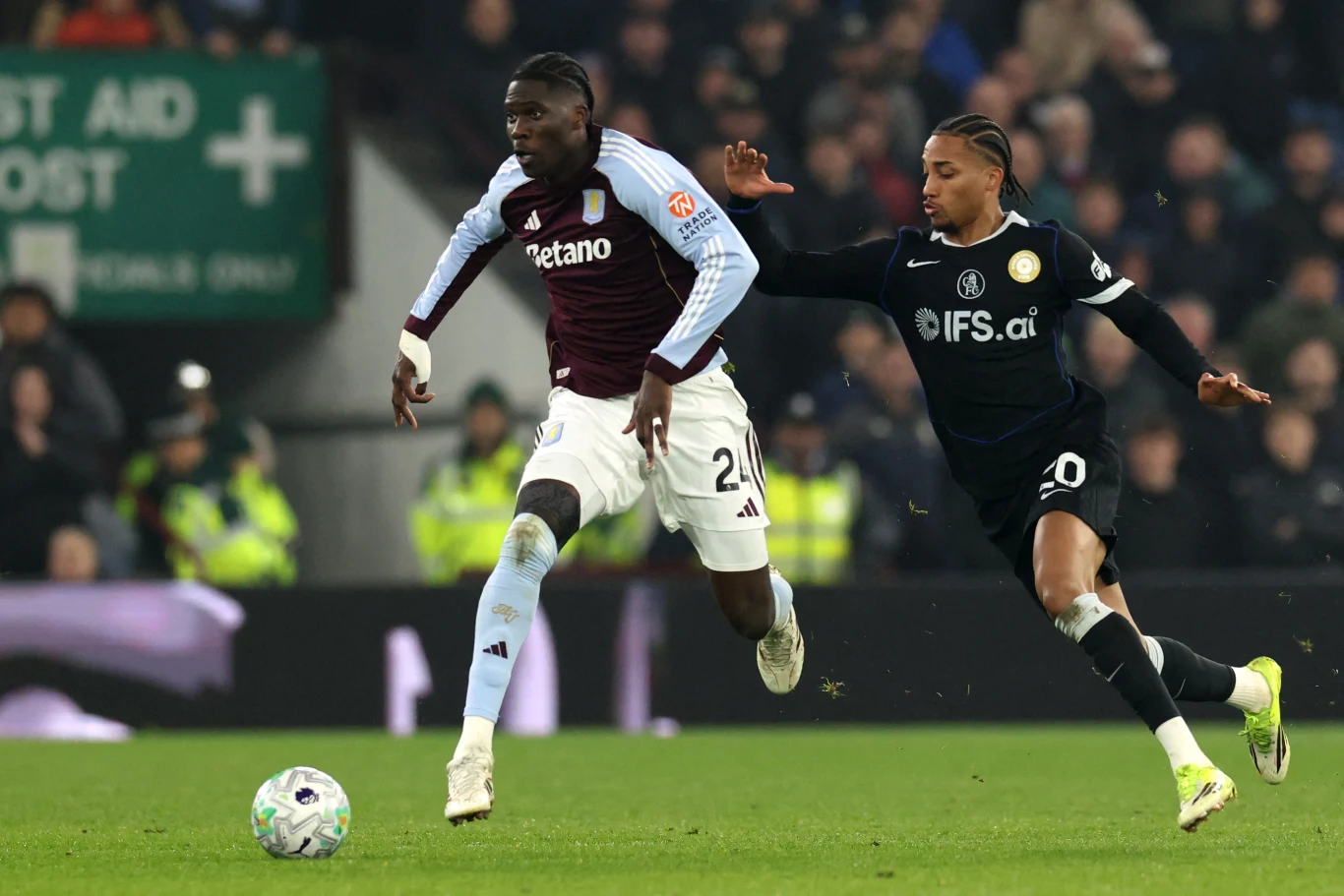 Dwóch piłkarzy w trakcie dynamicznej akcji na boisku – zawodnik w bordowo-błękitnej koszulce biegnie z piłką, a rywal w czarnym stroju próbuje go dogonić, w tle widoczna publiczność oraz służby porządkowe.