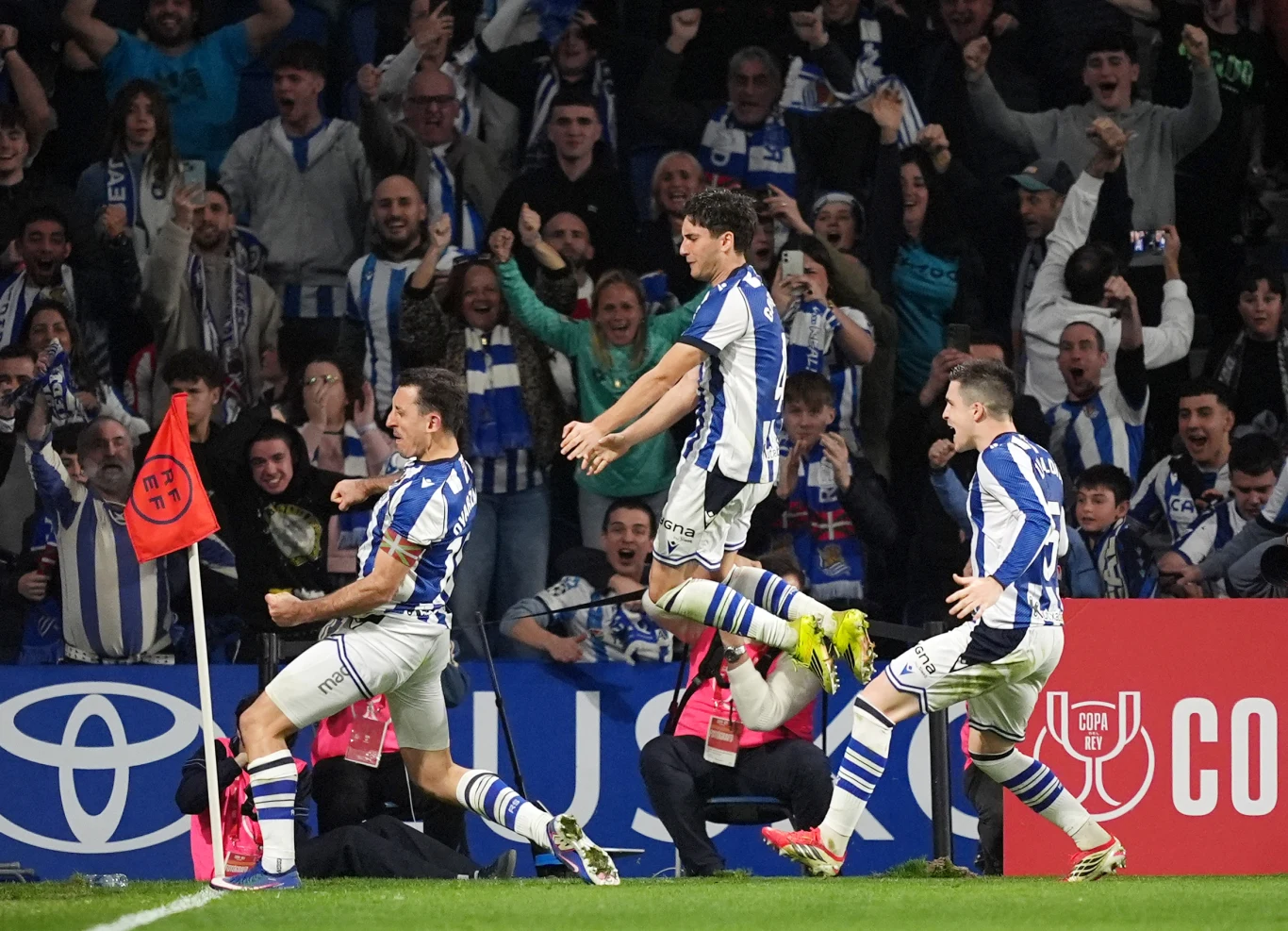 Trzech piłkarzy w niebiesko-białych strojach biegnie w kierunku narożnika boiska, świętując zdobycie gola, a za nimi tłum kibiców w euforii, podnoszących ręce i wyrażających radość.