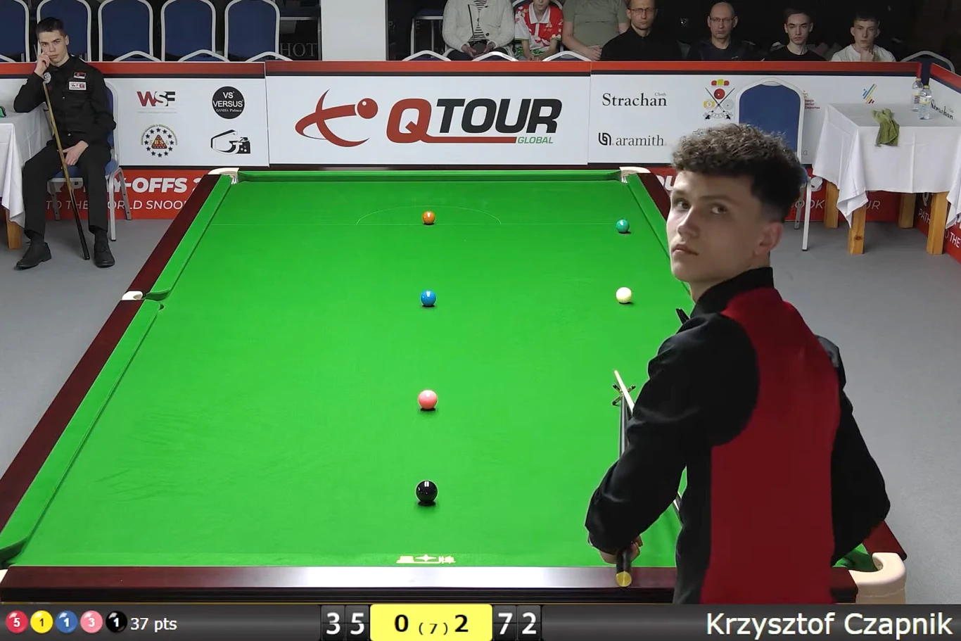 Zawodnik w czarno-czerwonym stroju snookerowym stoi przy zielonym stole z kijem w ręku; na stole rozmieszczone są bile snookerowe, a w tle widać sędziów oraz publiczność uważnie obserwującą rozgrywkę.