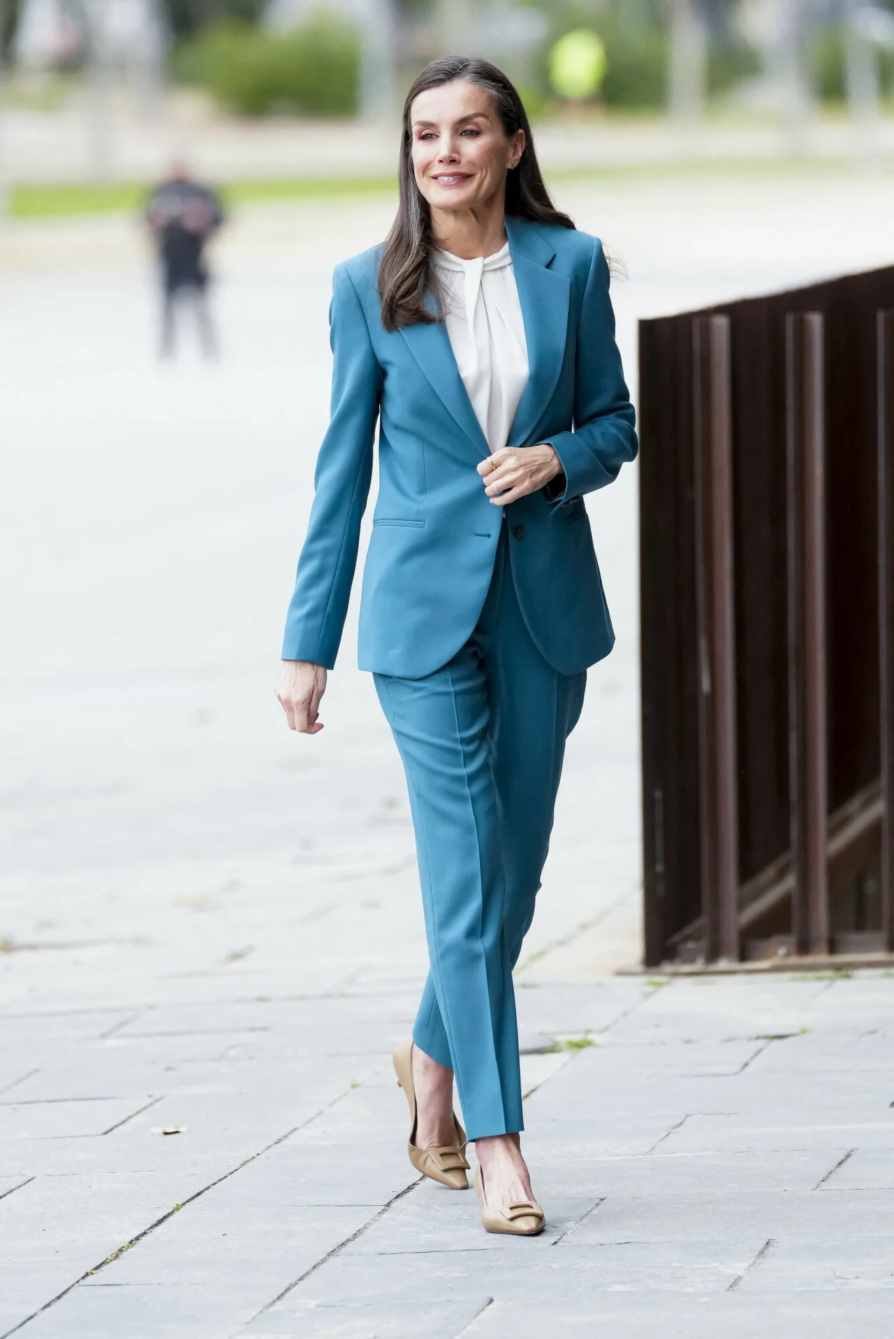 Kobieta ubrana w elegancki niebieski garnitur i białą bluzkę idzie chodnikiem, mając na sobie jasne buty na obcasie, z uśmiechem na twarzy i rozpuszczonymi włosami.