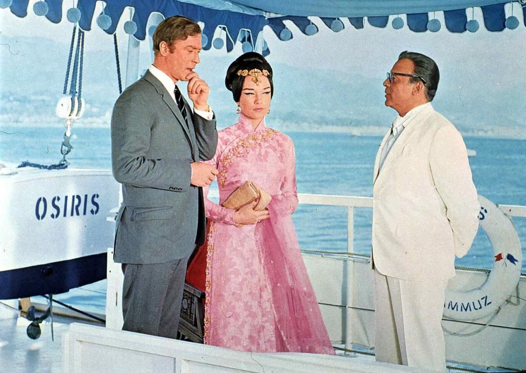 Trzy elegancko ubrane osoby stoją na pokładzie statku; mężczyzna w szarym garniturze i kobieta w różowej, tradycyjnej sukni z kopertówką naprzeciw starszego mężczyzny w białym garniturze, w tle widoczne koła ratunkowe oraz nazwa statku Osiris.