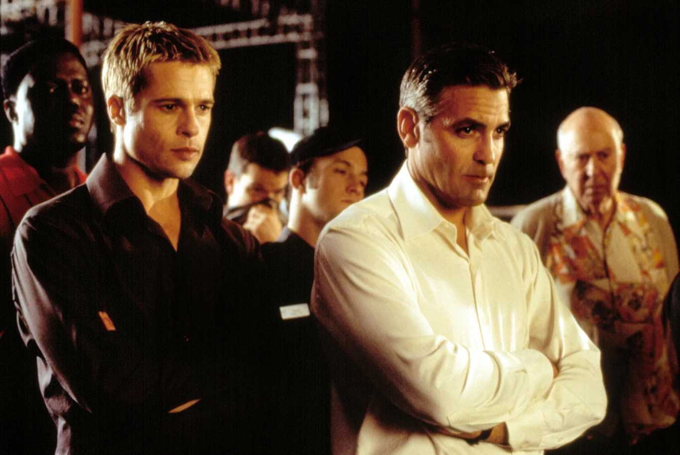 Dwie skupione osoby w eleganckich koszulach stoją w pierwszym planie z rękami skrzyżowanymi na piersiach, otoczone przez kilka innych postaci w tle, wyrażających zainteresowanie i koncentrację.