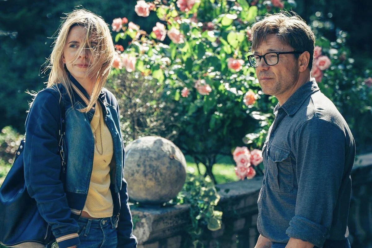 Kobieta w granatowej kurtce i mężczyzna w okularach stoją razem w ogrodzie pełnym różowych kwiatów, oboje patrzą w kierunku aparatu, a na pierwszym planie znajduje się kamienna kula na murku.