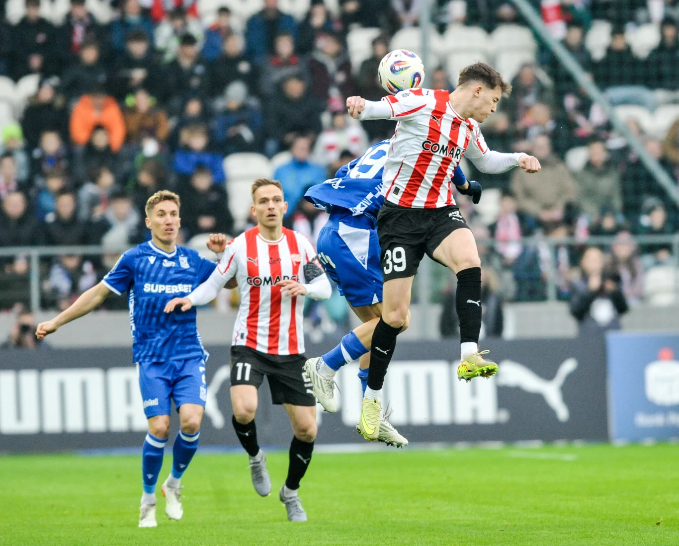 Dwóch piłkarzy walczy o piłkę głową podczas meczu piłkarskiego; zawodnik w biało-czerwonej koszulce wyskakuje wyżej niż rywal w niebieskim stroju, na drugim planie widać kibiców na trybunach.